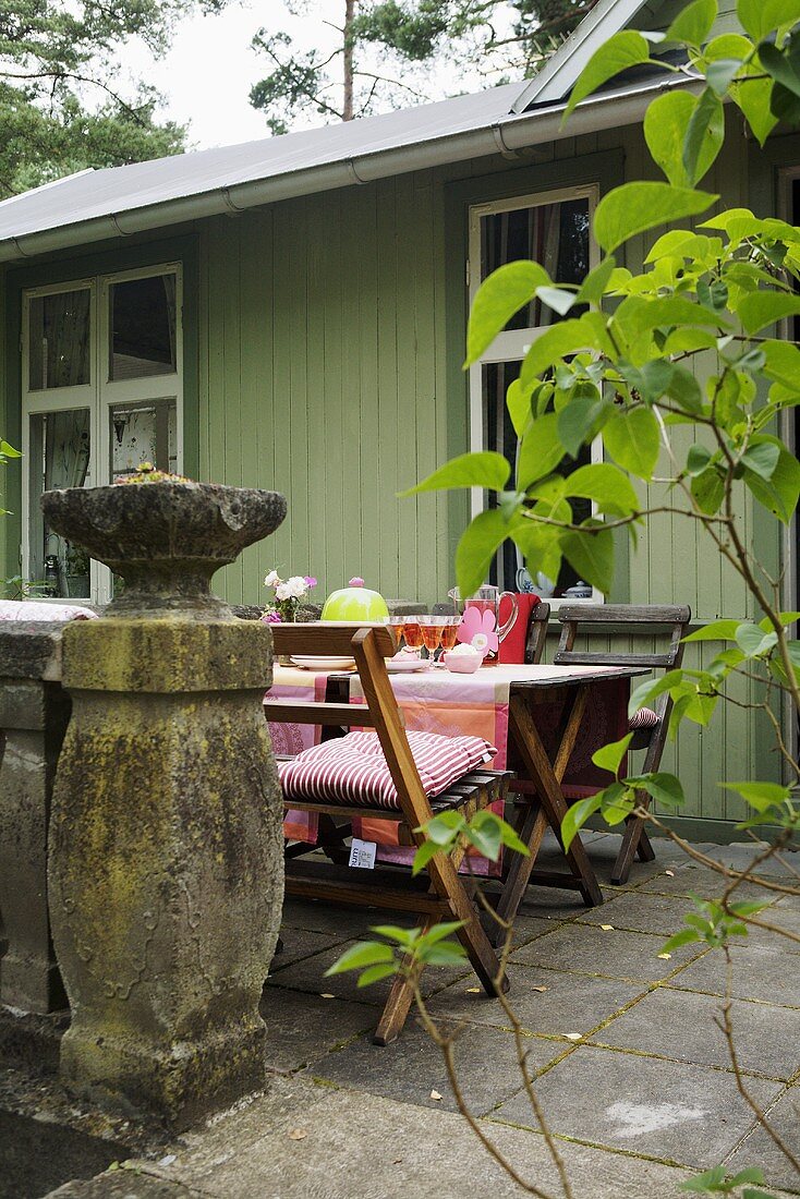 Gedeckter Tisch auf Steinterrasse vor grünem Holzhaus