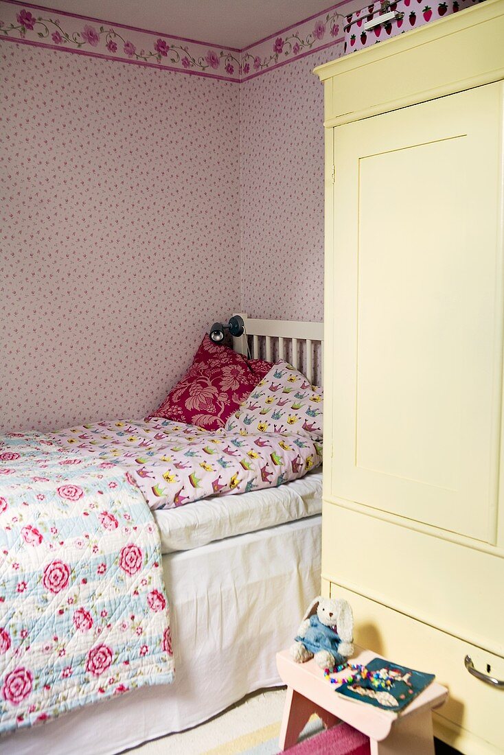 A child's bedroom with a bed, a cupboard and pink floral wallpaper on the wall