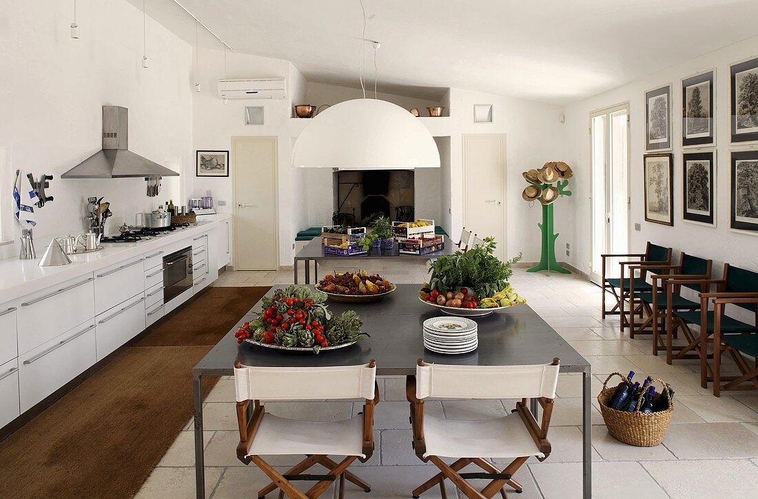 Küche mit Essplatz - Platten mit Gemüse auf grauem Metalltisch und Holzklappstühle mit weißem Stoff auf weißem Fliesenboden