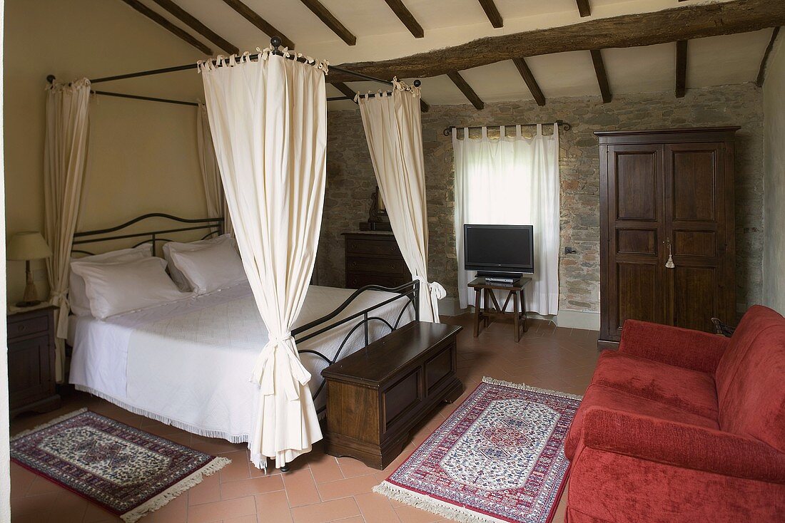 Holzbalkendecke im rustikalen Schlafraum mit Himmelbett und Sofa mit rotem Samtbezug