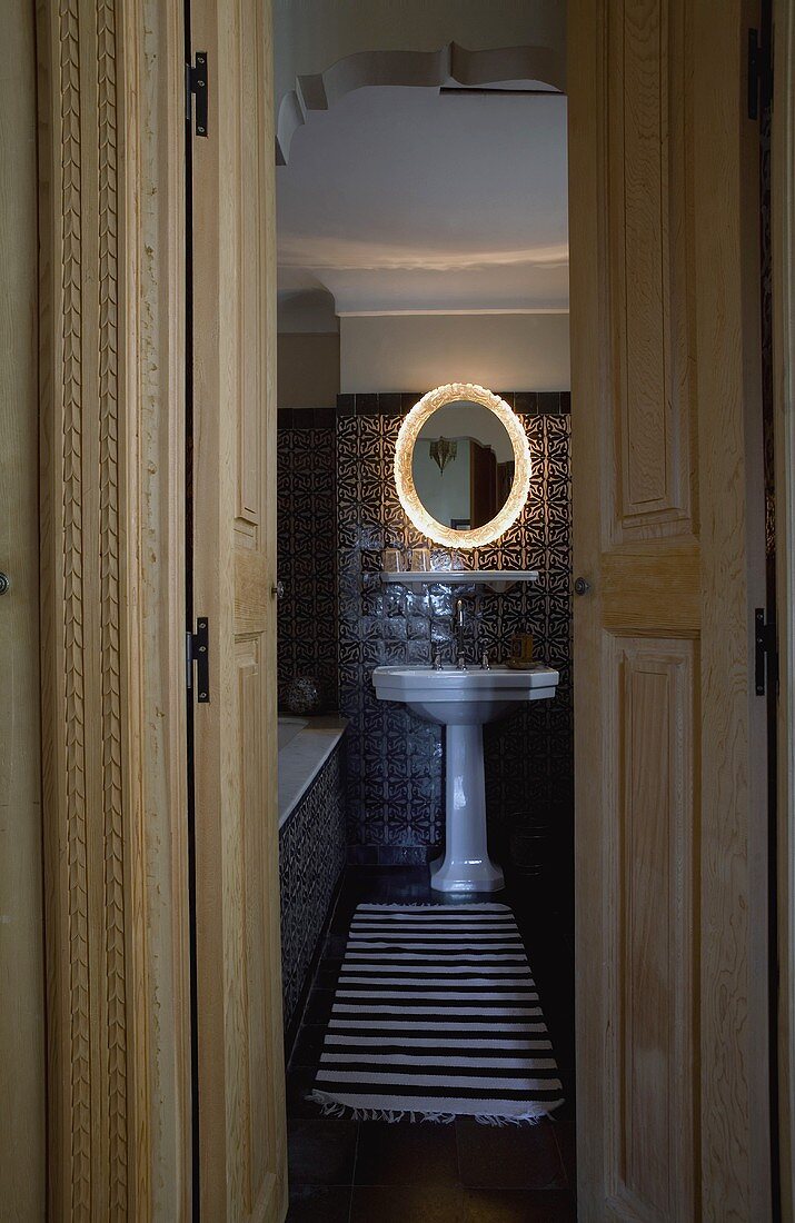 Open door with a view into a bathroom with a white pedestal sink with a lighted mirror