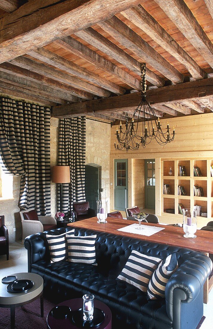 Open living-dining room in a country home with a wooden beam ceiling, striped floor length curtains hanging at the windows