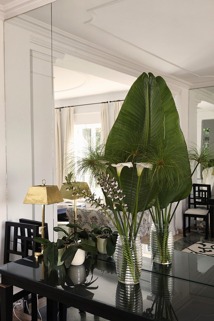 Palm frond and tropical flowers in vases on a shiny, black wall table in front of a mirrored wall