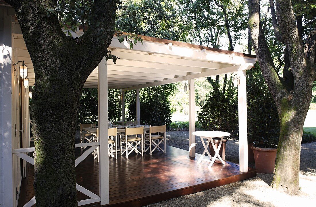 Trees in front of white wooden terrace with posts and a patio table on a raised wooden platform