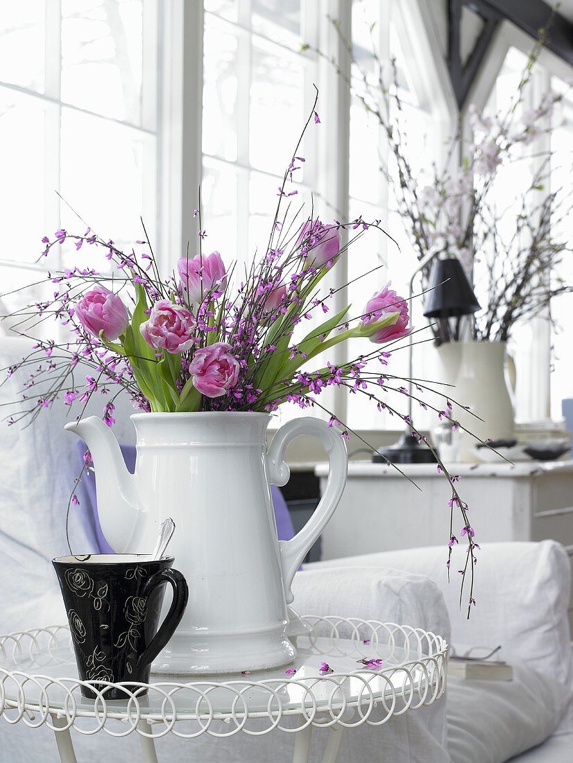 Strauss mit pinkfarbenen Tulpen und blühenden Zweigen in weisser Porzellankanne auf Beistelltisch