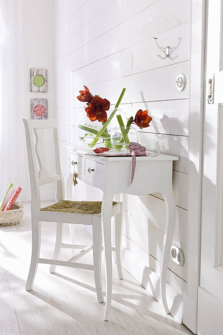 Weisser Wandtisch mit Stuhl vor weissvertäfelter Wand und roten Amaryllisblumen in Glasvase