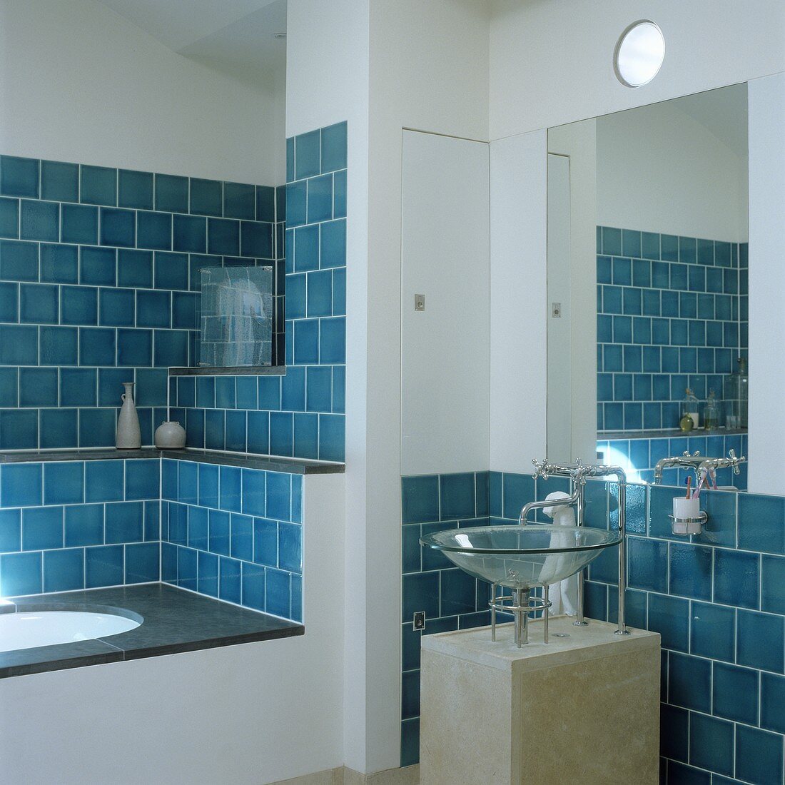 Blue wall tiles in a niche with a built-in bathtub and a glass sink on a concrete pedestal with a mirror above