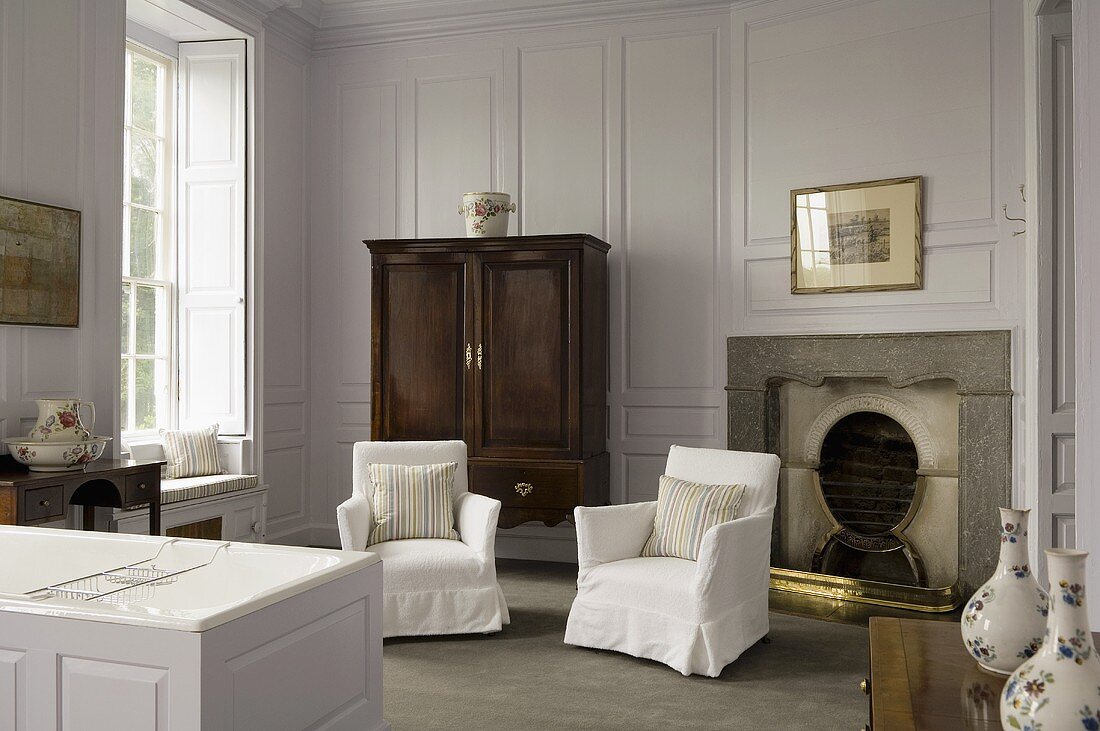 An elegantly designed country house-style bathroom with white armchairs in front of a fireplace and lilac-painted walls
