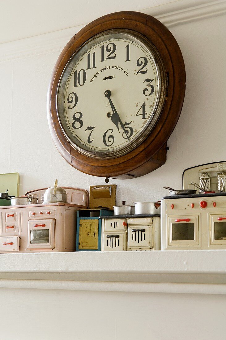 An antique wall clock in a round wooden frame above a shelf with a dolls kitchen