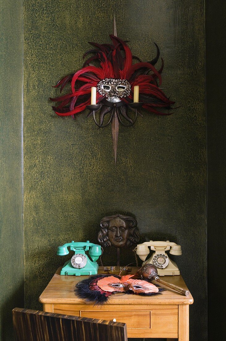 Wandtischchen aus hellem Holz mit alten Telefonapparaten und Kerzenleuchter mit Maske vor dunkler Wand