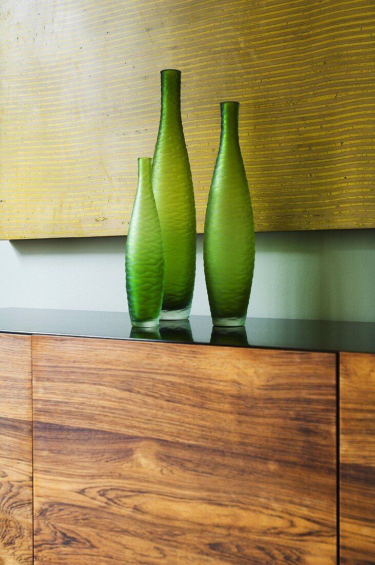 Green vases on a wooden side board in front of a yellow picture