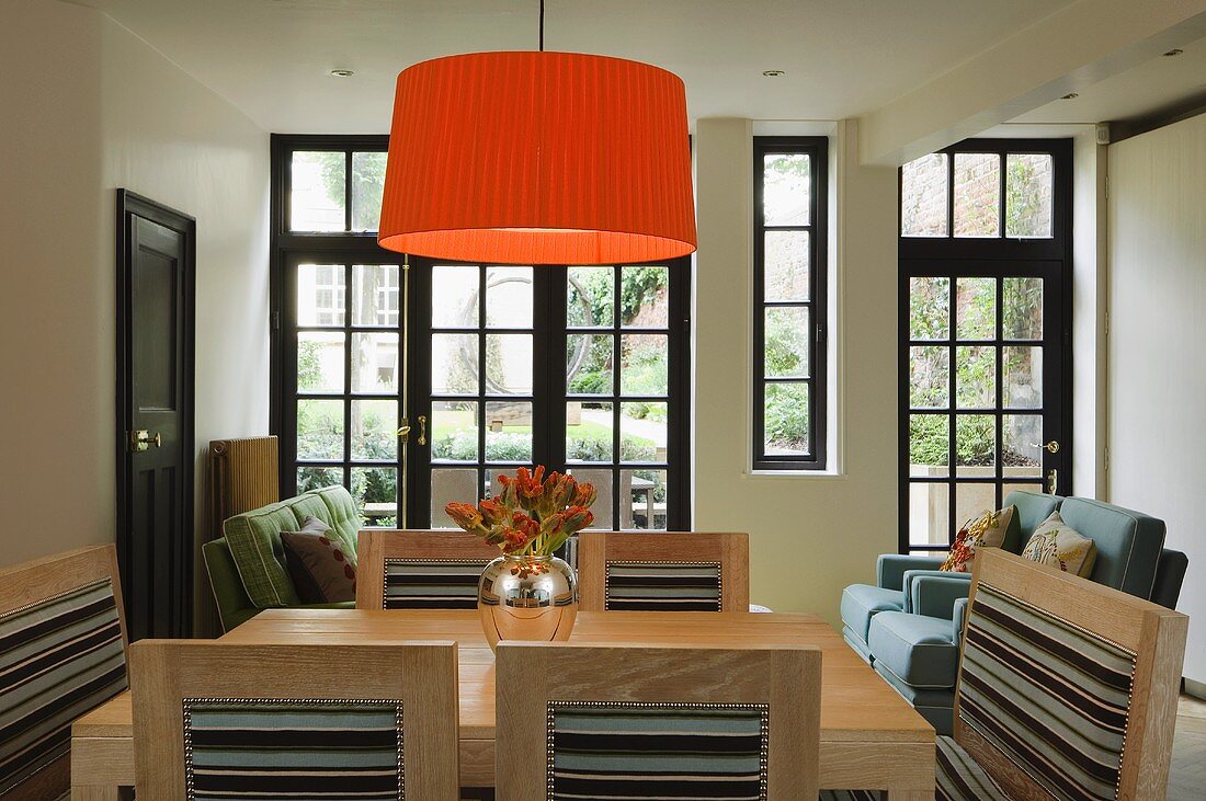 A pleated orange lampshade above a light coloured dining table and chairs in front of a bank of windows with black frames