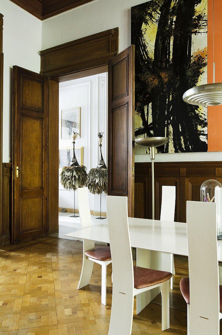 A wood panelled dining room with a white designer table and white designer chairs and a view through the open double doors onto the sculptures in the hallway