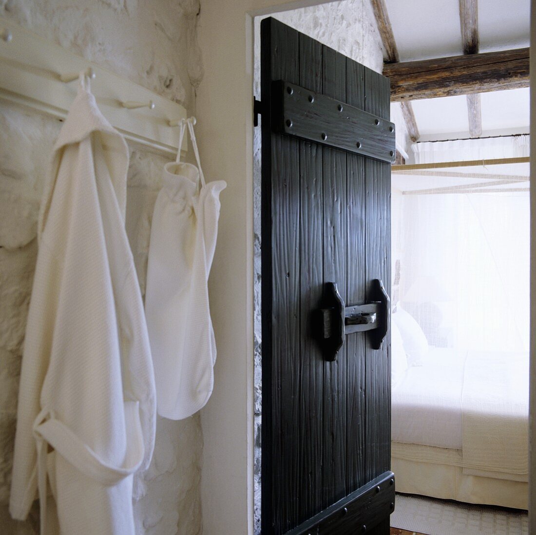 A rustic country house - a bathrobe on a hook next to an open, grey wooden door with a view of a four-poster bed