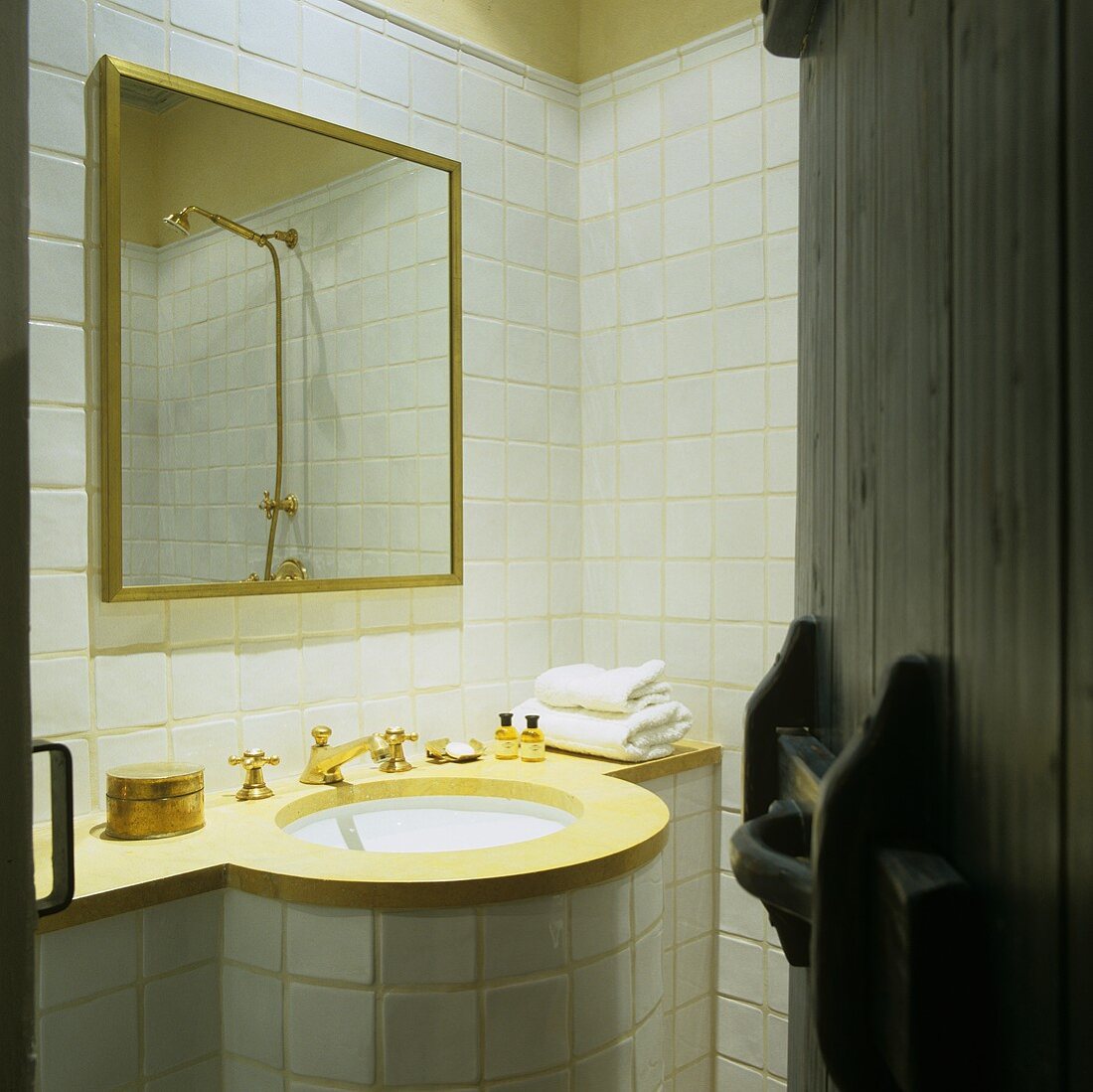 A view into a white tiled bathroom with a round basin with brass taps and a gold-framed mirror