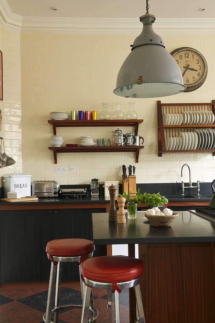 An open-plan kitchen in a country house - a metal lamp over a kitchen counter with red upholstered bar stools and wooden shelves on the wall