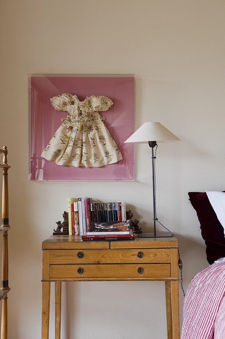 A table lamp with a white shade on a light wood bedside table with a framed doll's dress hanging on the wall