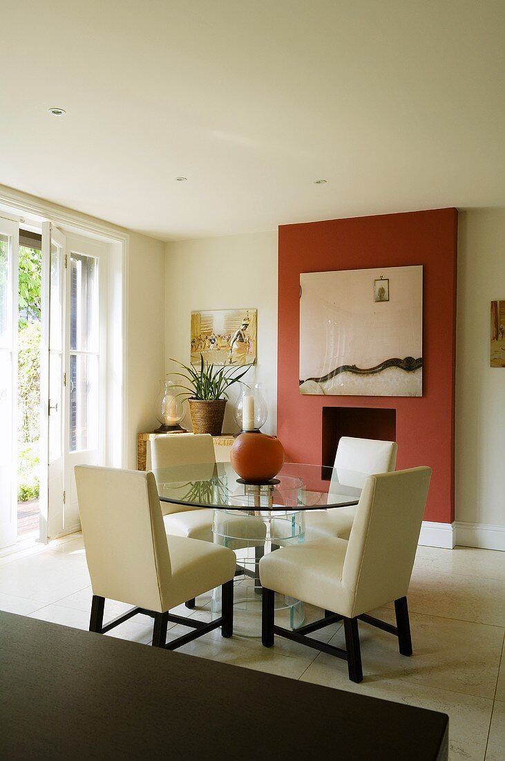 Eleganter Wohnraum mit gepolsterten weissen Stühlen und rundem Glastisch vor Kamin in rotgetönter Wandscheibe