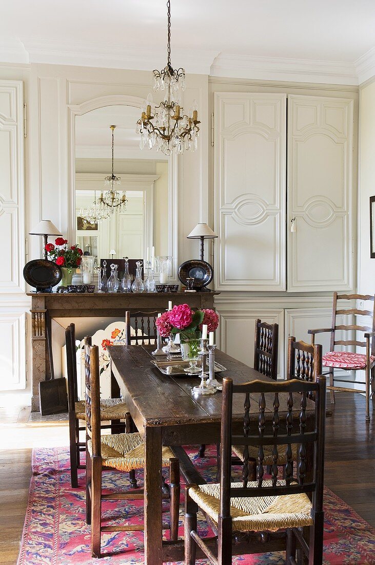 A rustic dining table with antique, country house-style chairs and light grey wood panelling with built in cupboards