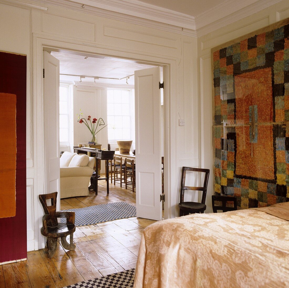 Moderner Wandteppich vor weiss vertäfelter Wand im Schlafraum mit Blick durch offene Flügeltür in Wohn- und Essraum