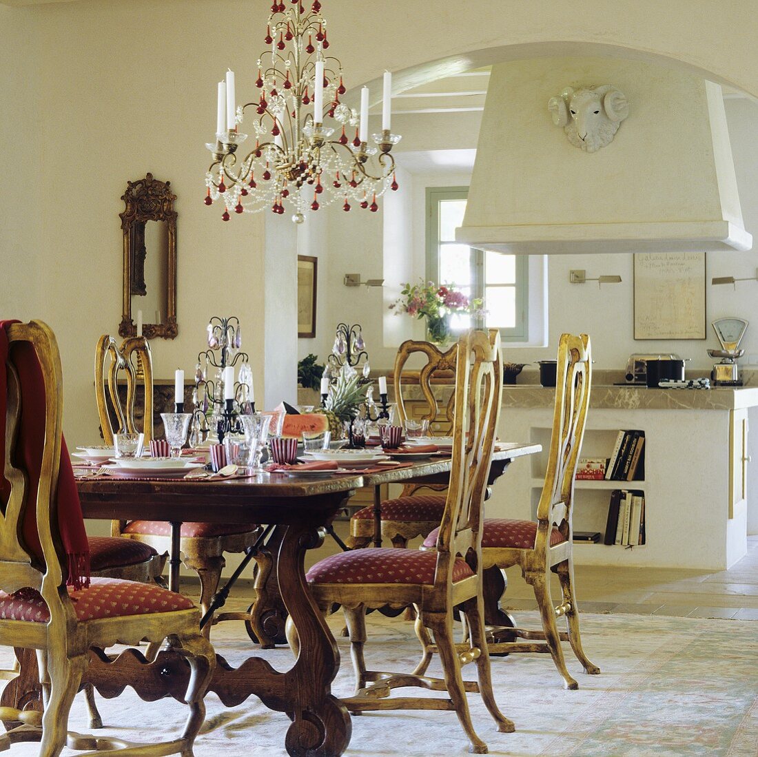 A laid table set with upholstered country house chairs and a chandelier with an open Mediterranean-style kitchen in the background