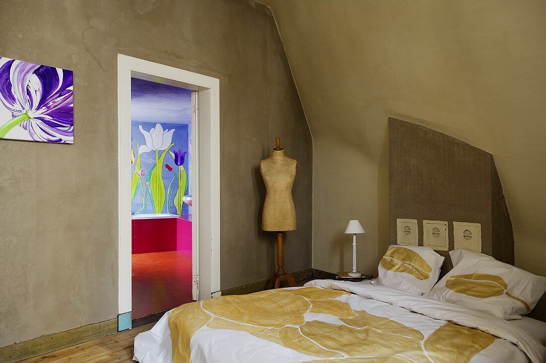 A grey bedroom with a double bed and a tailor's dummy next to a door opening with view into a colourful bathroom
