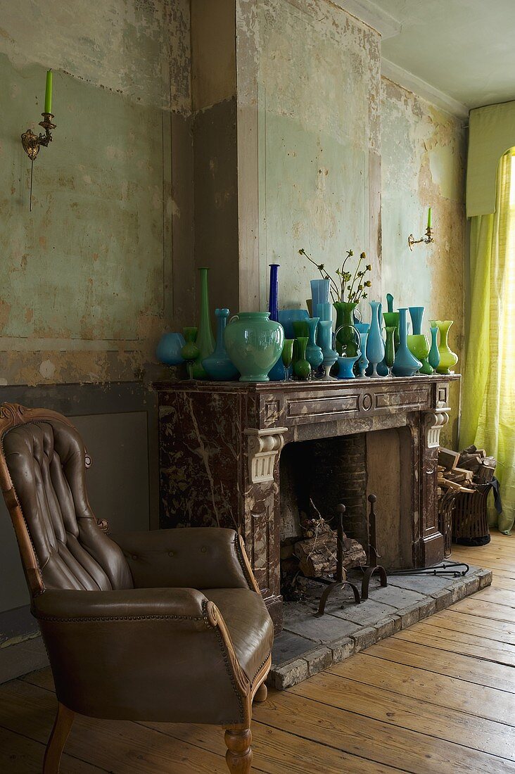 An antique leather armchair next to a fireplace with a collection of blue-green vases on the mantelpiece of an old fashioned living room