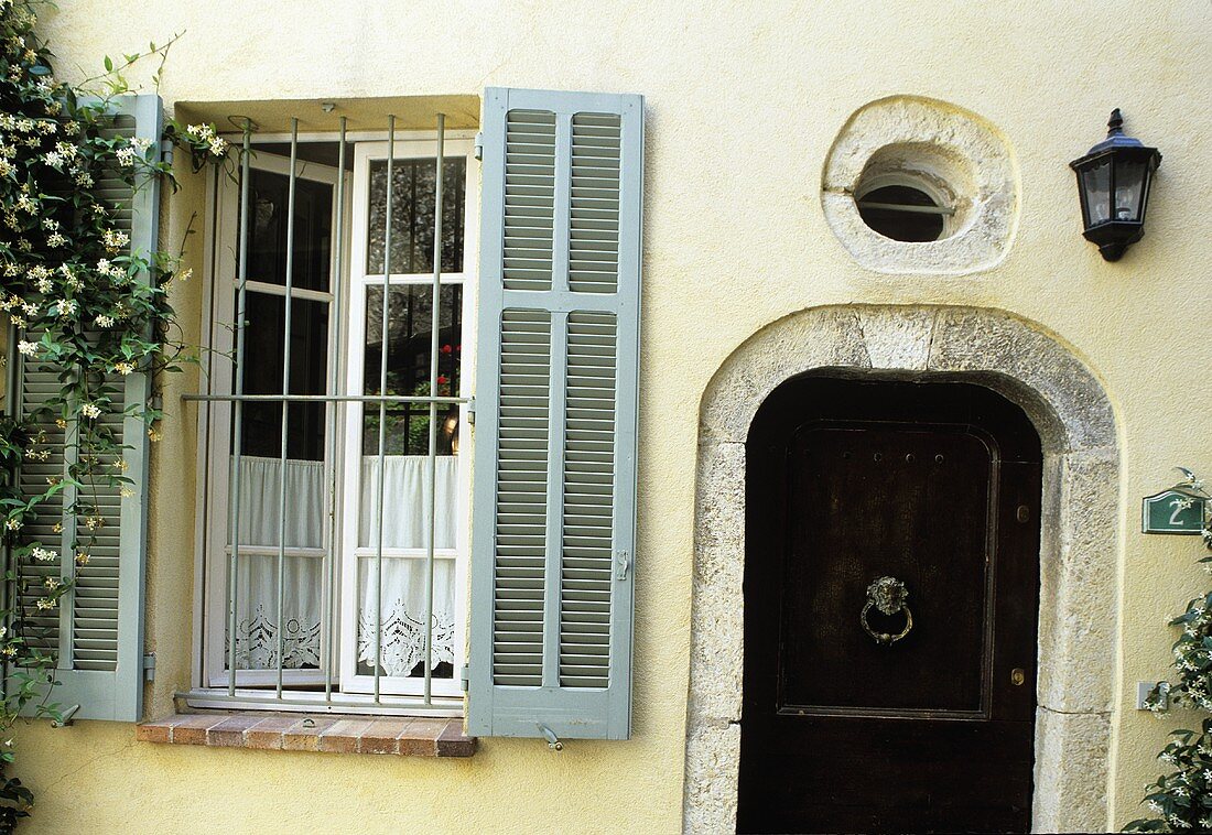 Vergittertes Fenster mit grauem Holzladen und Haustür aus dunklem Holz mit Steineinfassung und Oberlicht in hellgelb getönter Fassade