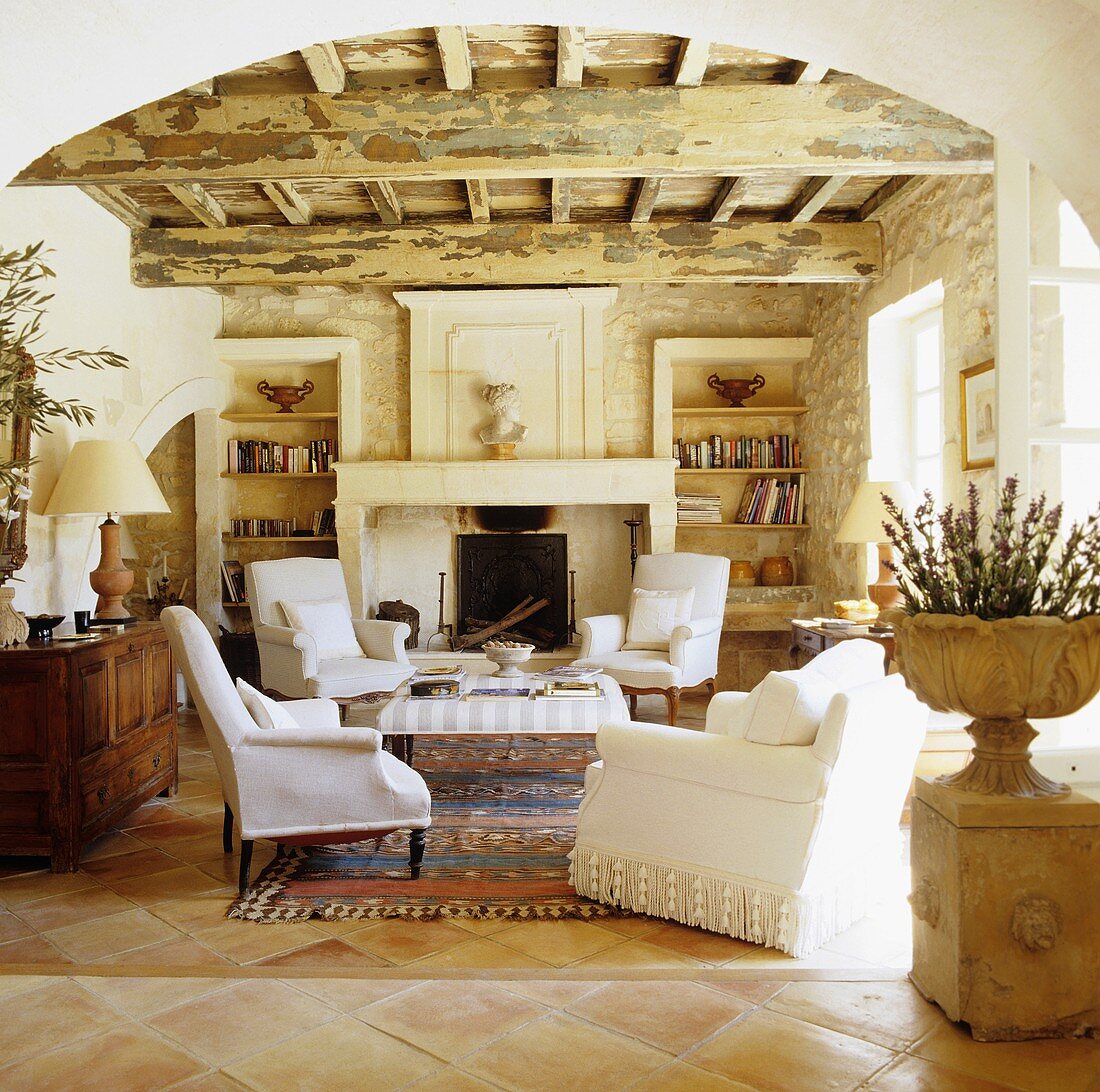 A view through a rounded archway in a Mediterranean living room with a rustic wood beam ceiling and white armchairs in front of a natural stone fireplace