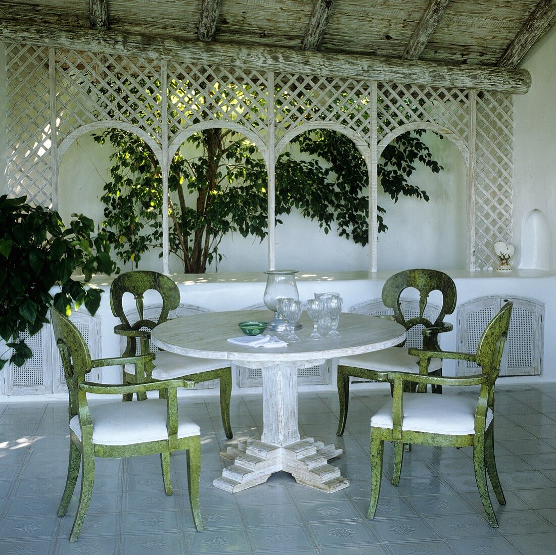 Green curved wooden chairs around a round table and wooden lattice wall with rounded arches on a veranda