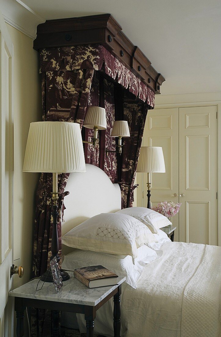 Table and wall lamps next to and above a double bed with patterned canopy and white bed linen