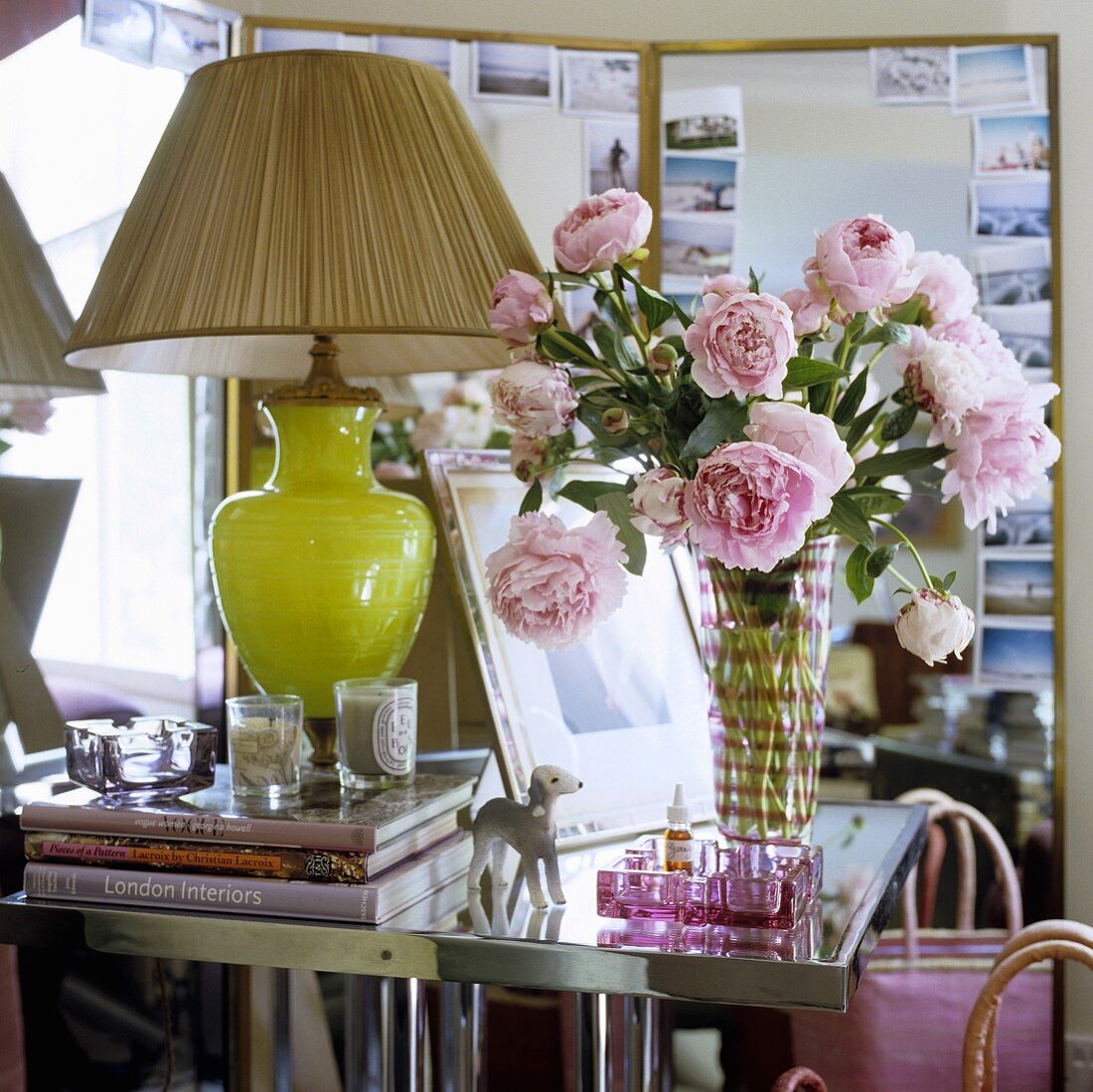A table lamp with a green glass base and a pleated shade next to a bunch of roses in a glass vase on a side table