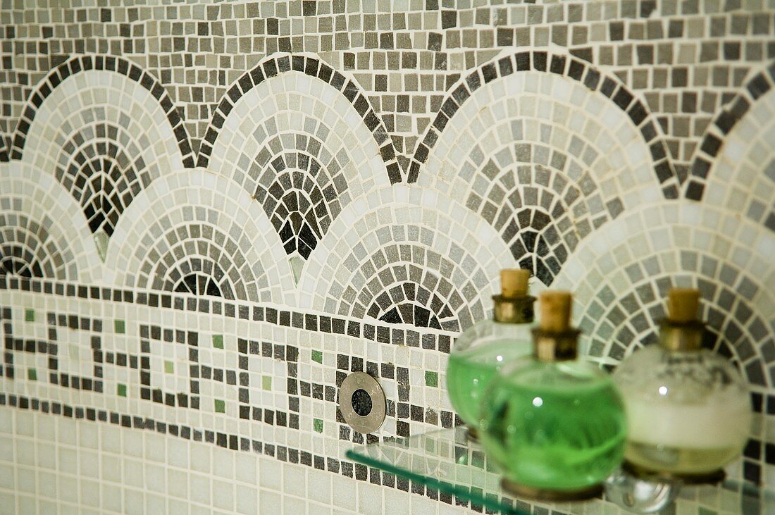Glass bottles on a glass shelf in front of a circular design made of mosaic tiles on a wall
