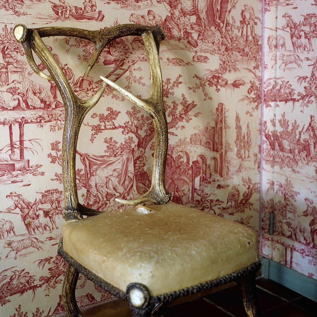 Gepolsterter Stuhl mit verarbeitetem Hirschgeweih vor gemusterter Tapete auf Wand