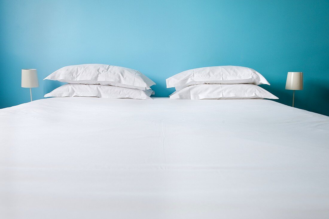 Bett mit weisser Bettwäsche und Kopfkissen vor blauer Wand