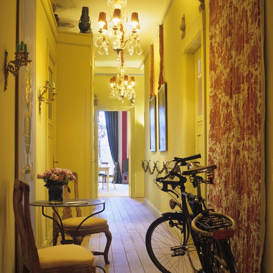 A bicycle, wooden chairs and an occasional table in a narrow hallway with yellow walls and chandeliers