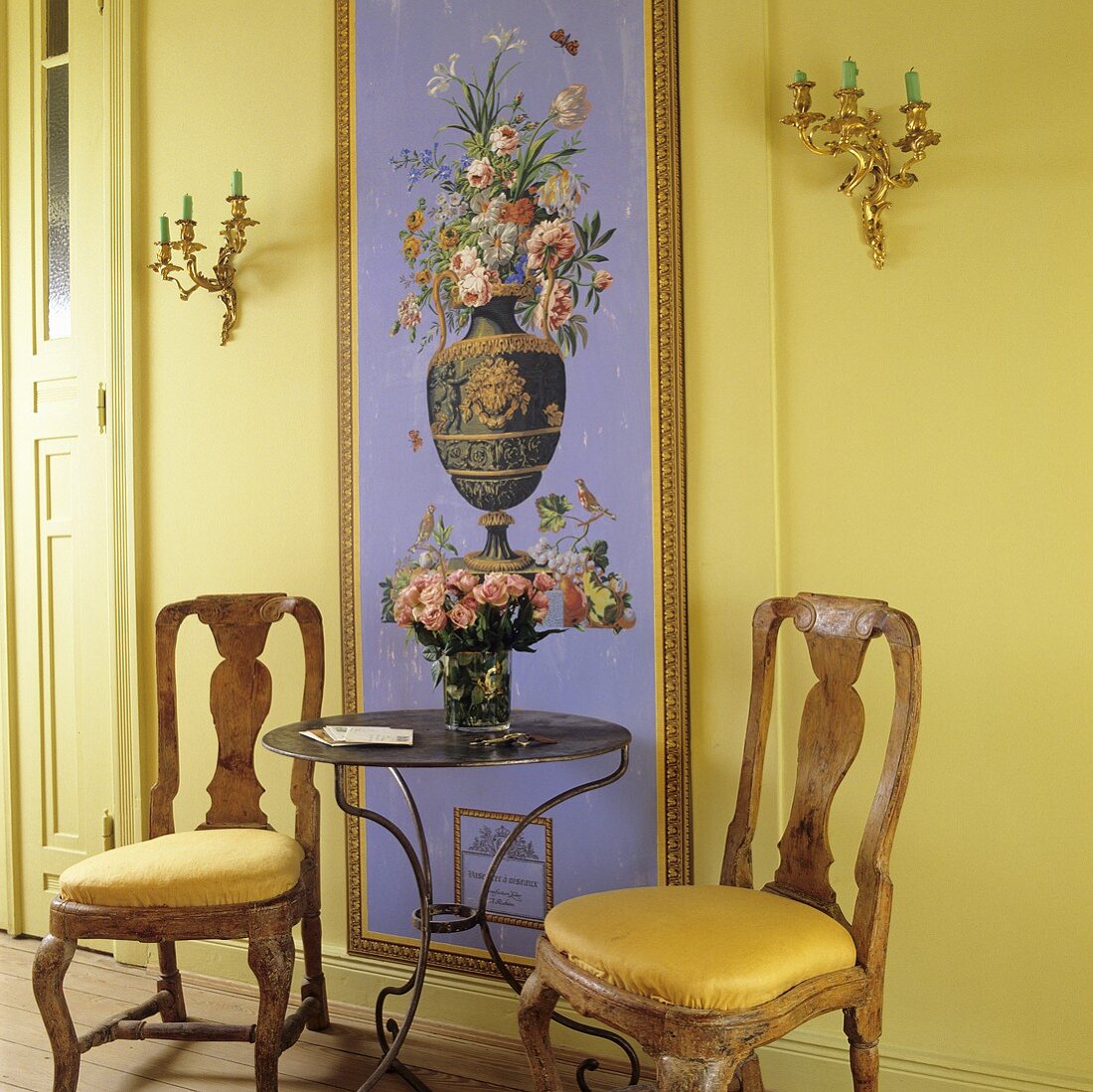 Antique wooden chairs and a side table in a hallway front of a yellow wall with a frame floral picture
