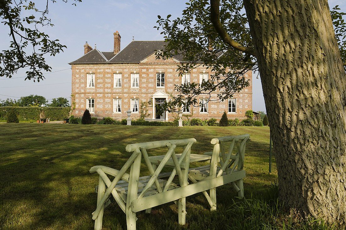 A wooden bench under a tree and a view across a garden to a French manor house