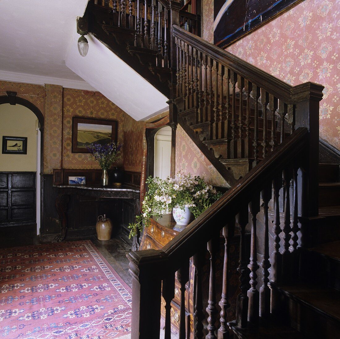 Vorraum eines Landhauses mit dunkler Holztreppe und Muster auf roter Tapete