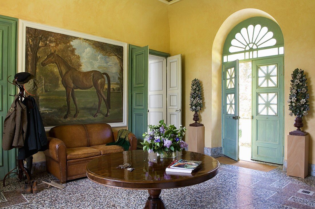 A leather sofa and a wardrobe in front of a large picture of a horse in a sunny yellow hallway in a country house