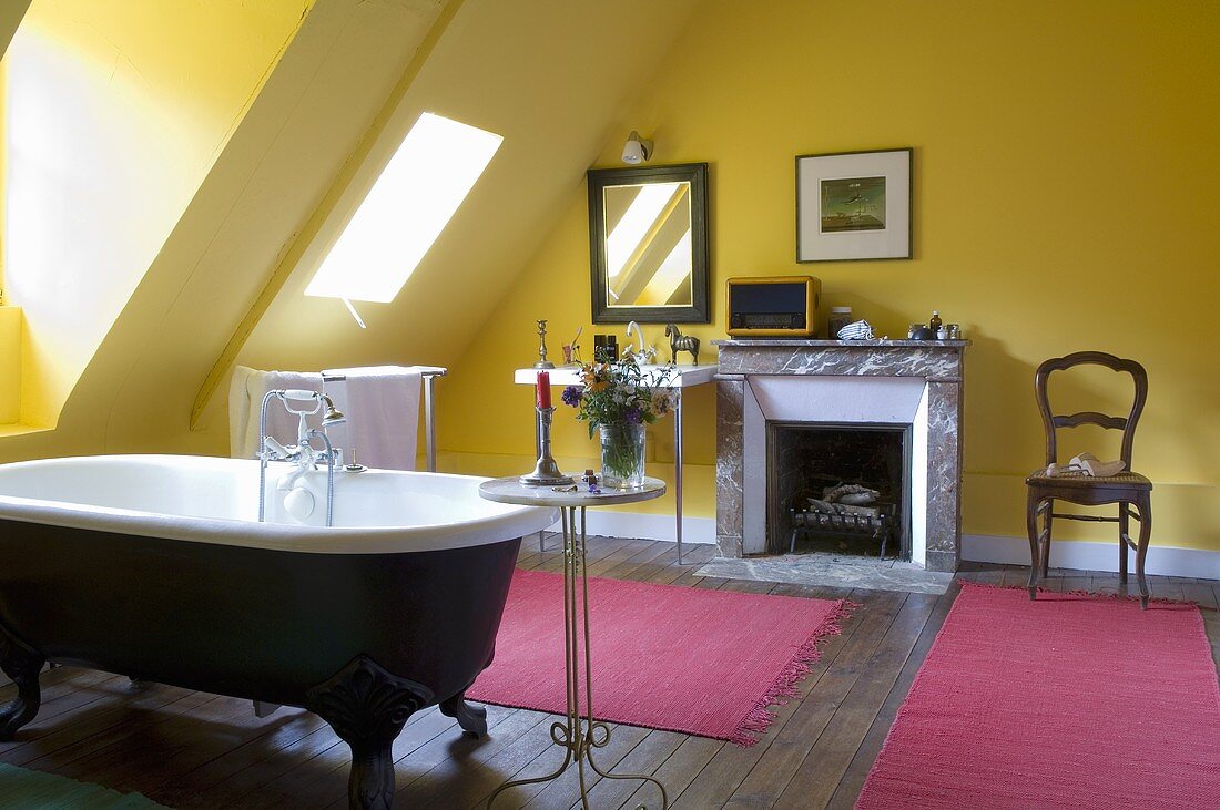 Gelbgetöntes Bad mit Kamin unter Dachschräge und pinkfarbene Badematten vor freistehender antiker Badewanne