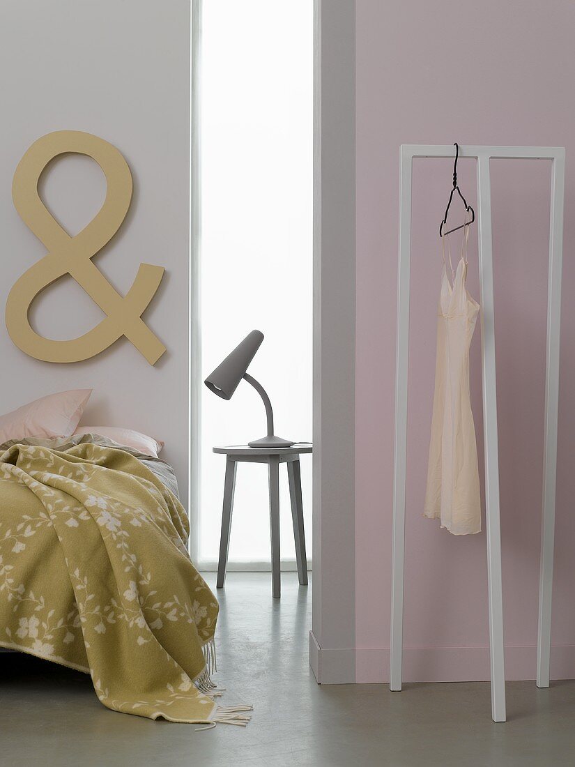 A lamp on a bedside table between a bed and a room divider with a clothes rack
