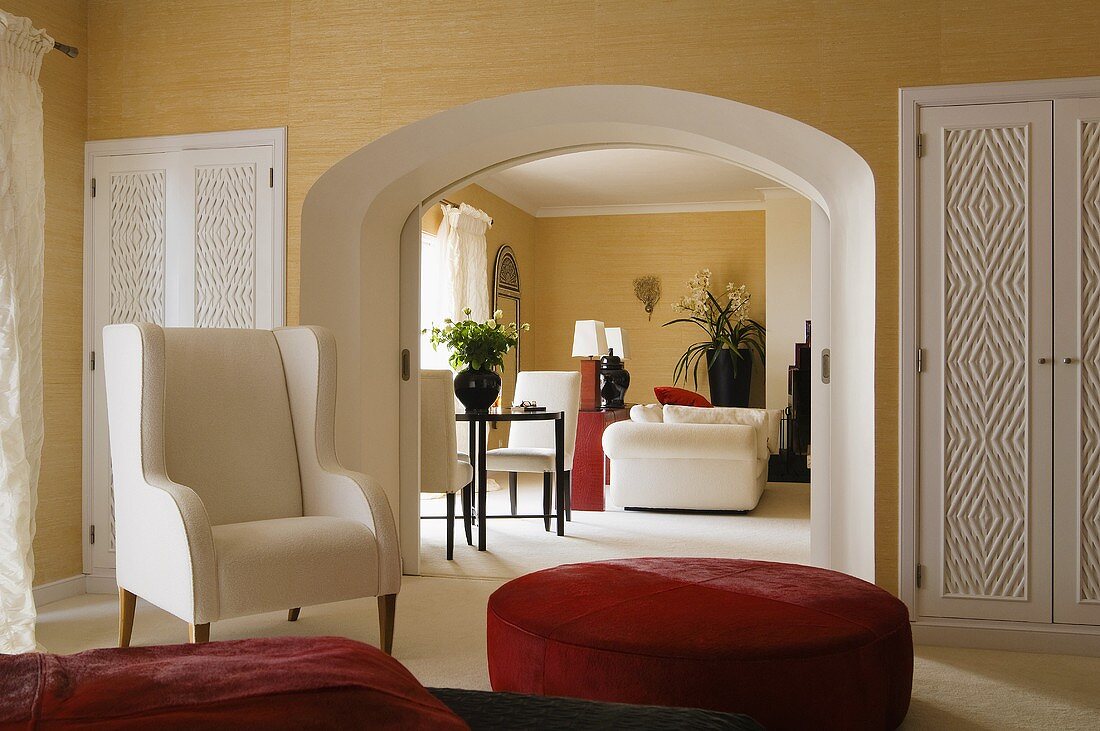 A white armchair and a red upholstered stool in a bedroom with a view through an arched doorway into a living room