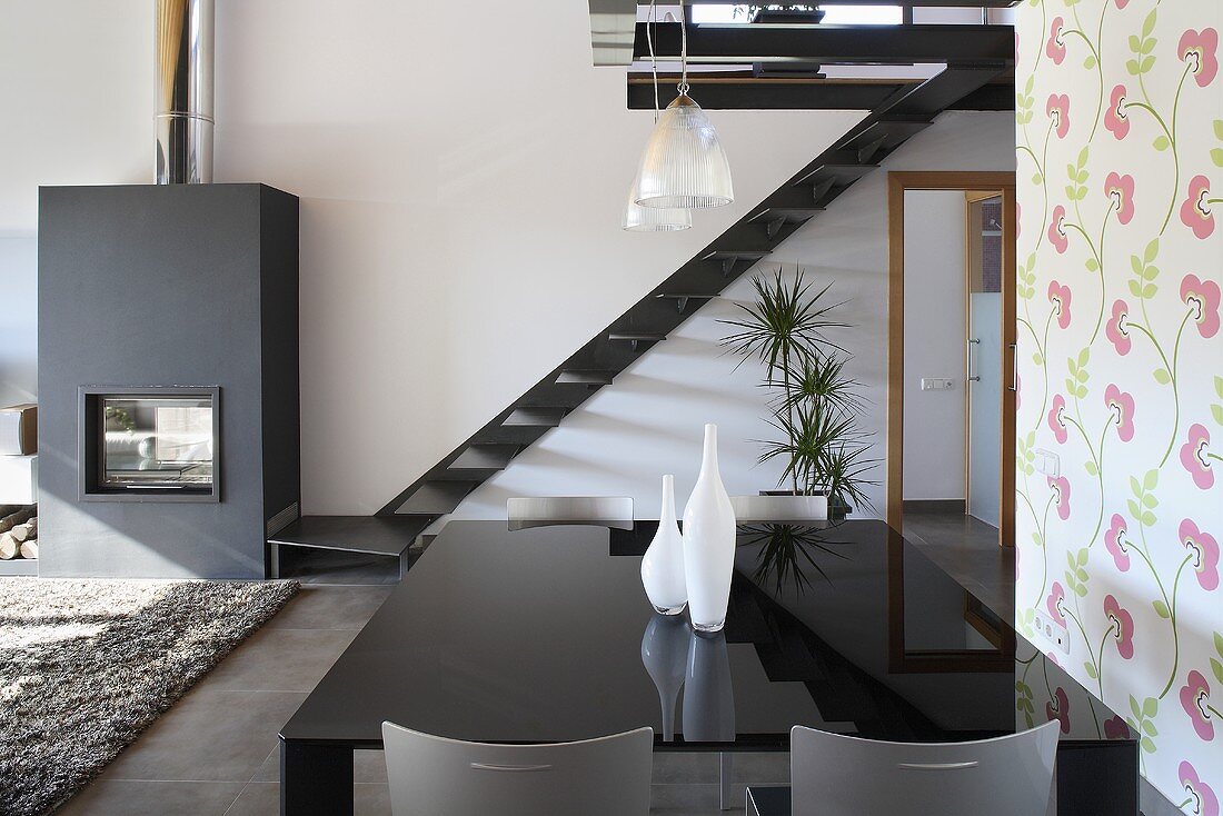 An open living-room-cum-dining-room with a black shiny table in front of a flight of stairs and a fireplace