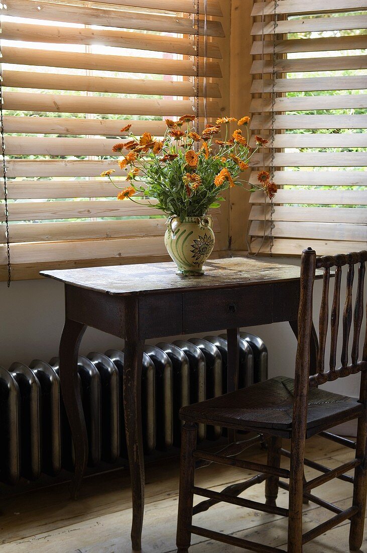 A bunch of garden flowers on a simple wooden davenport with a chair in front of wooden blinds