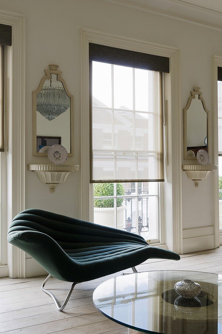 A curved chaise lounge in front of a floor-to-ceiling window with a blind and antique brackets on the walls with mirrors above them