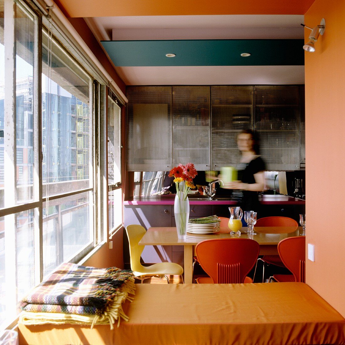 Durchgang mit Blick auf offene Küche, Frau steht am Essplatz mit farbigen Stühlen vor Glasfront