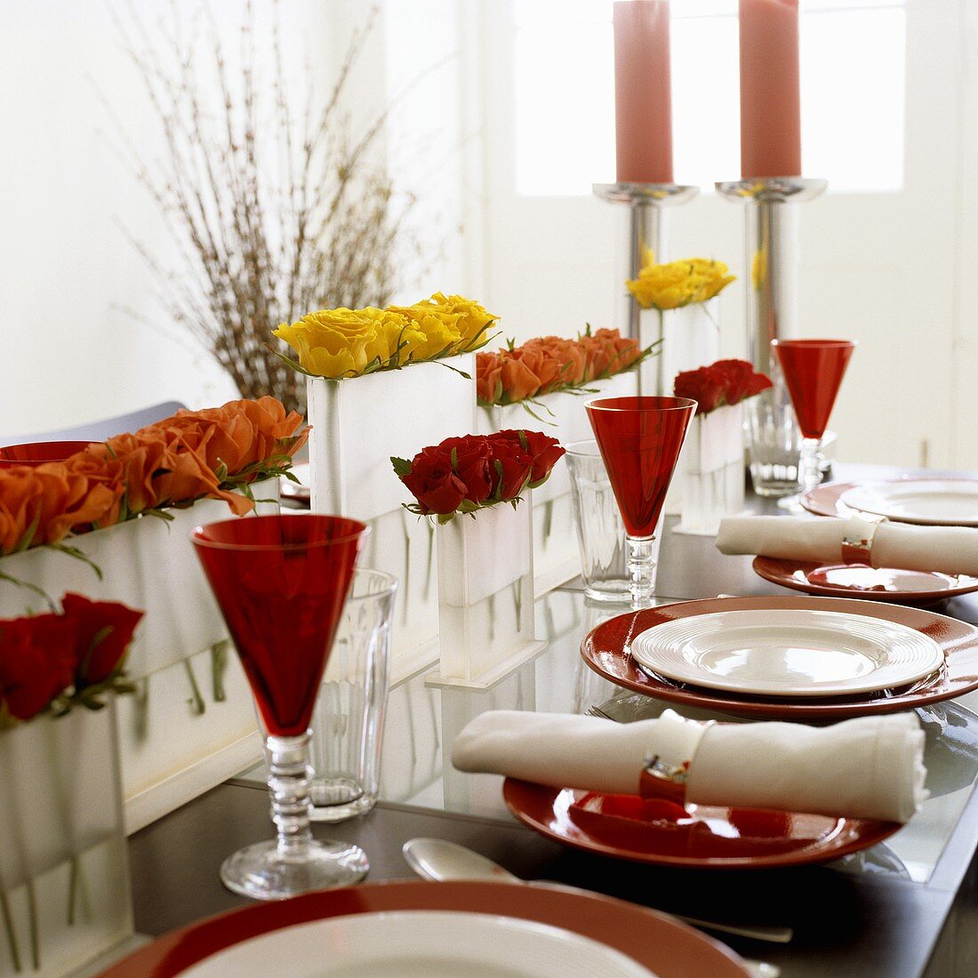 A laid table - red and white place settings with coloured stem glasses and roses in vases