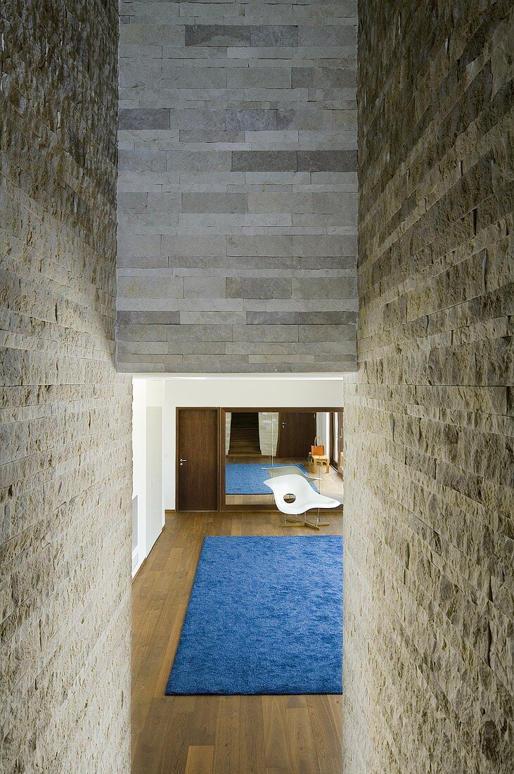 A natural stone wall with a view through a narrow opening into a living room with a wooden floor and a blue rug