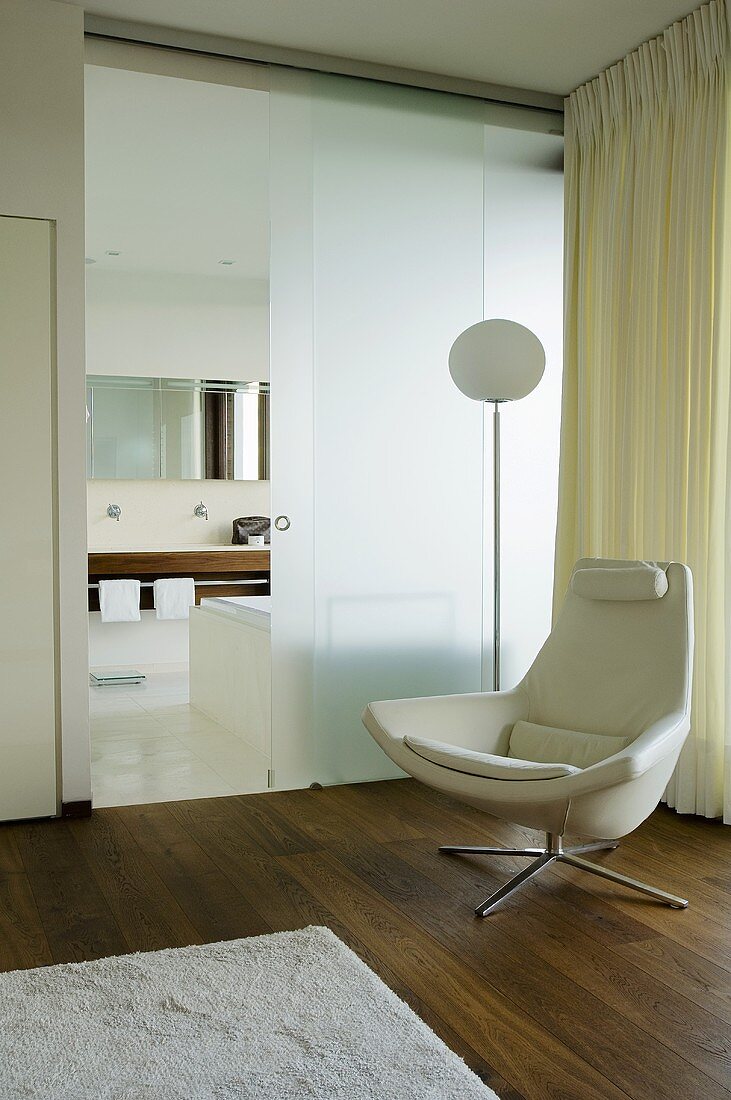 Designerledersessel auf Nussholzboden vor opaker Glaswand mit offener Schiebetür und Blick ins Bad